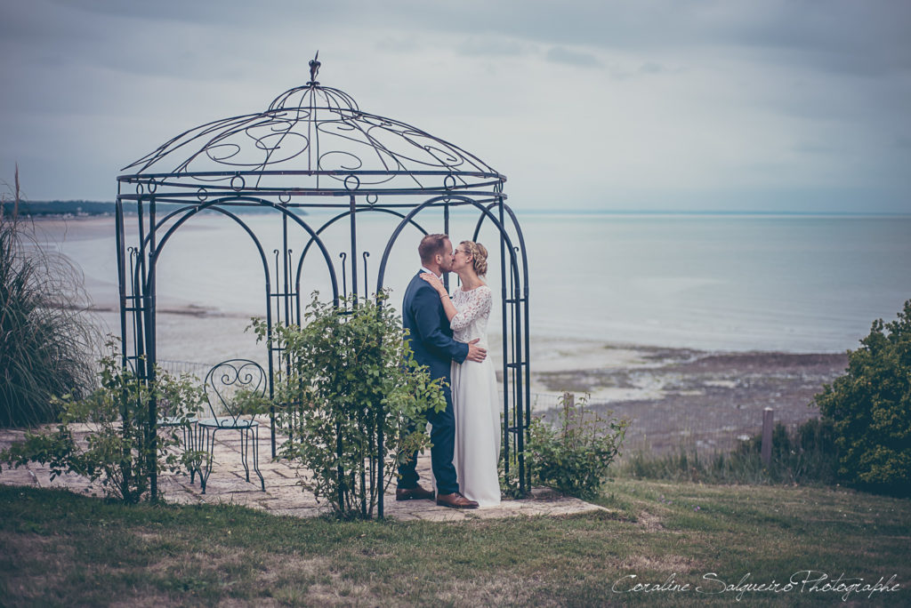 couple de mariés qui s'embrasse sou sune gloriette avec la mer en toile de fond